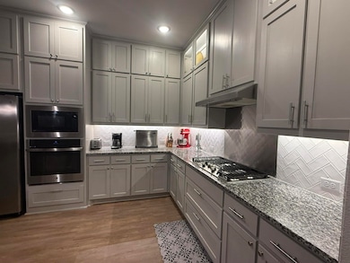 Kitchen featuring light stone counters, gray cabinets, decorative backsplash, appliances with stainless steel finishes, and under cabinet range hood