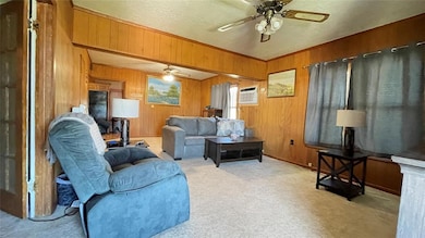 Carpeted living room featuring ceiling fan, wood walls, and an AC wall unit
