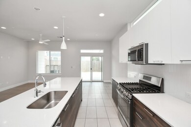 Modern kitchen with sleek white countertops, dark cabinetry, and stainless steel appliances. Features an island with a sink and ample natural light from the rear glass door.