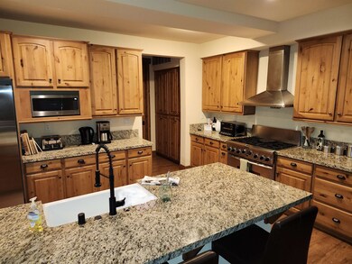 Kitchen featuring wall chimney range hood, light stone countertops, stainless steel appliances, dark wood-style flooring, and brown cabinets