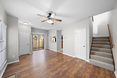This inviting living space features hardwood floors, a ceiling fan, and neutral walls. It includes access to a patio through sliding glass doors, a staircase leading upstairs, and connects to the kitchen area.