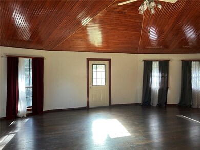Spacious living room with a high, wooden vaulted ceiling and dark vinyl plank floors. It features large windows with curtains, allowing natural light, and a central door. Ceiling fan and light fixture add functionality.