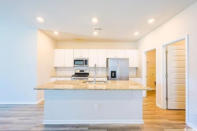 Kitchen with white cabinetry, light stone counters, appliances with stainless steel finishes, a center island with sink, and light wood-style flooring