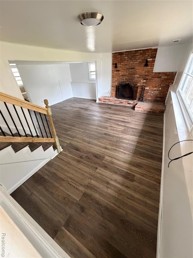 Unfurnished living room featuring dark wood finished floors, plenty of natural light, a brick fireplace, and stairway