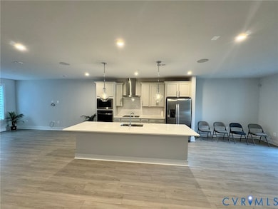 Kitchen featuring open floor plan, an island with sink, wall chimney range hood, pendant lighting, and white cabinetry