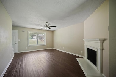 Unfurnished living room with dark wood finished floors, a textured ceiling, a ceiling fan, a fireplace with raised hearth, and baseboards