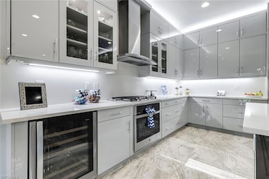 light gray cabinets, wall chimney range hood, wine cooler, stainless steel appliances, and glass insert cabinets