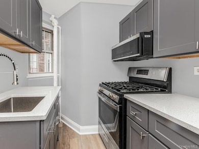 Kitchen with gray cabinetry, appliances with stainless steel finishes, light wood-type flooring, and light stone counters