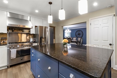 Kitchen with wall chimney range hood, appliances with stainless steel finishes, blue cabinets, a chandelier, and white cabinets