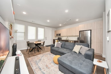 Modern, open-concept living space with a sleek kitchen, featuring stainless steel appliances and light wood cabinetry. The living area has a comfortable sectional sofa and a stylish dining table by large windows, creating a bright and inviting atmosphere.