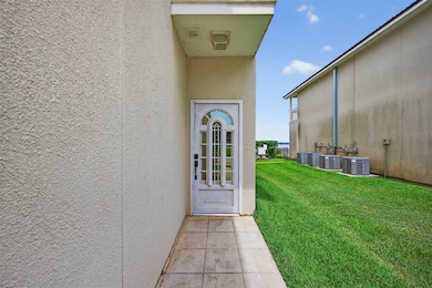 A narrow entryway leads to a white door with decorative glass, situated between two buildings. The area is paved and opens to a grassy side yard with air conditioning units.