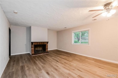 Unfurnished living room featuring a textured wall, wood finished floors, a brick fireplace, ceiling fan, and a textured ceiling