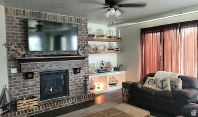 Living room with wood finished floors, a textured ceiling, ceiling fan, and a brick fireplace