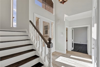 Bright and welcoming entryway featuring a staircase with dark wood steps and white railings. Large windows allow natural light to fill the space, leading to an adjoining room with dark flooring and French doors.