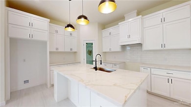 Kitchen featuring light stone counters, white cabinetry, pendant lighting, a center island with sink, and backsplash