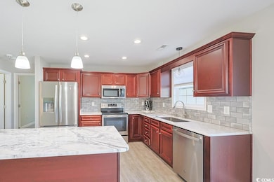 Kitchen with reddish brown cabinets, appliances with stainless steel finishes, light stone countertops, decorative light fixtures, and backsplash