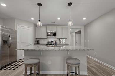 Kitchen featuring stainless steel appliances, light stone countertops, a center island with sink, light wood finished floors, and hanging light fixtures