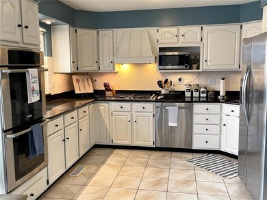 Gas cooktop with double ovens, gleaming stainless appliances, custom downdraft hood cabinet, raised panel cabinets with black pulls and hinges, granite counters.