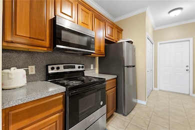 Kitchen featuring stainless steel appliances, brown cabinetry, crown molding, light tile patterned floors, and decorative backsplash