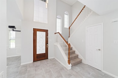 Bright and welcoming entryway featuring a wooden front door with decorative glass, tiled flooring, and a staircase with wooden railings. High ceilings and large windows enhance natural light.
