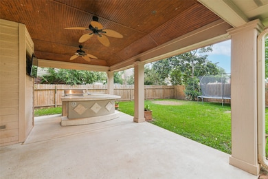 Spacious covered patio with a wood-paneled ceiling, featuring two ceiling fans and a built-in outdoor kitchen with a grill. The area overlooks a fenced backyard (trampoline will be removed)  ideal for entertaining and family activities.