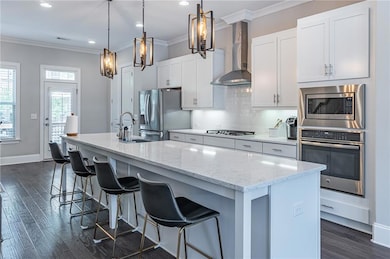 Kitchen with appliances with stainless steel finishes, wall chimney range hood, a sink, a kitchen island with sink, and ornamental molding