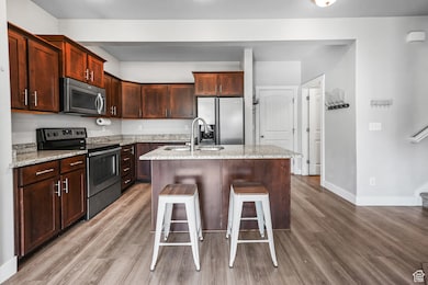 Kitchen featuring appliances with stainless steel finishes, a center island with sink, light stone countertops, light wood-type flooring, and dark brown cabinets