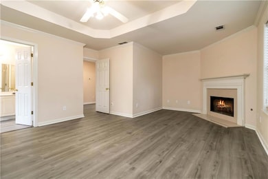 Unfurnished living room featuring ceiling fan, a raised ceiling, wood finished floors, a warm lit fireplace, and crown molding