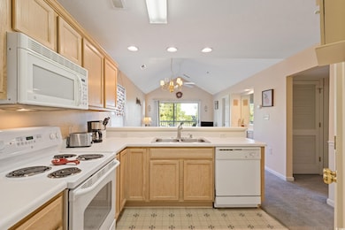 Kitchen with light brown cabinetry, white appliances, vaulted ceiling, a chandelier, and a peninsula