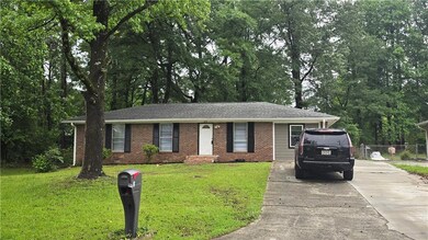 Single story home featuring driveway, brick siding, roof with shingles, and view of wooded area