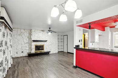 Kitchen featuring a ceiling fan, a stone fireplace, dark countertops, wood finished floors, and open floor plan
