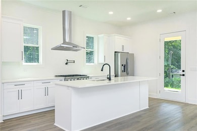 Kitchen featuring wall chimney exhaust hood, stainless steel refrigerator with ice dispenser, a kitchen island with sink, wood finished floors, and white cabinetry