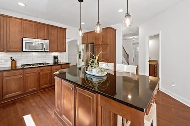 Kitchen featuring appliances with stainless steel finishes, dark wood-type flooring, decorative backsplash, an island with sink, and brown cabinetry