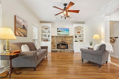 Living room featuring light wood finished floors, ornamental molding, built in features, a stone fireplace, and a ceiling fan