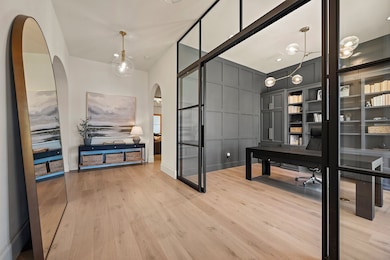 The office sits just off the entry, framed by sleek glass walls and rich paneling that give it a polished, modern feel. Arched walkways and warm wood flooring carry through from the foyer, setting the tone for the home’s thoughtful and cohesive design.