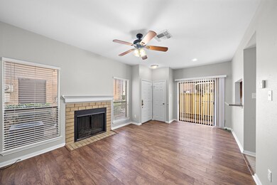 Cozy living room with a brick fireplace, vynil flooring, and ample natural light from large windows. Features a ceiling fan and sliding glass door access to an outdoor space. Ideal for comfortable living and entertaining.