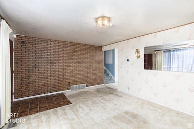 Carpeted empty room with wallpapered walls, stairway, and brick wall