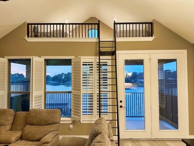 This inviting living room features vaulted ceilings and a cozy loft accessible by a ladder. Large windows and glass doors with shutters offer stunning waterfront views and natural light.