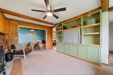 Living area with wood walls, a ceiling fan, light carpet, beam ceiling, and a fireplace
