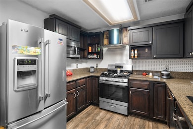 Kitchen featuring stainless steel appliances, open shelves, dark stone counters, wall chimney exhaust hood, and dark wood-style floors