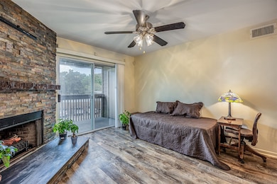 This cozy room features a stone fireplace, wood look flooring, and a ceiling fan. It opens to a balcony through sliding glass doors, providing natural light and a view.