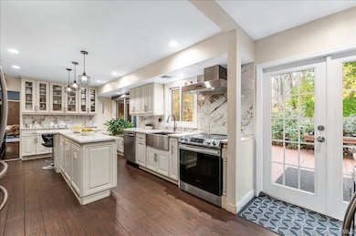 Kitchen featuring glass insert cabinets, stainless steel appliances, dark wood finished floors, wall chimney range hood, and pendant lighting