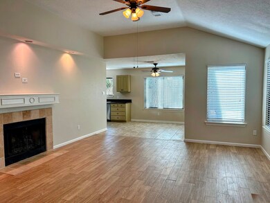 Bright, open-concept living area with wood flooring, a cozy fireplace, and ceiling fans. Large windows provide abundant natural light, and the space seamlessly connects to a tiled kitchen area.