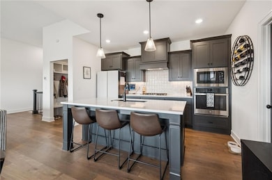 Kitchen featuring decorative backsplash, stainless steel appliances, a center island with sink, pendant light fixtures, and engineered hardwood floors