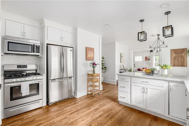 Kitchen featuring stainless steel appliances, white cabinets, decorative light fixtures, and light wood finished floors