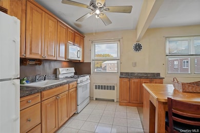 Kitchen featuring white appliances, dark countertops, light tile patterned floors, radiator, and brown cabinetry