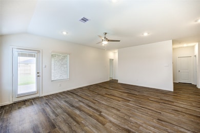 This spacious living area features modern wood flooring, neutral walls, recessed lighting, and a ceiling fan. It has a door with window blinds leading to the outside and ample natural light from a large window.