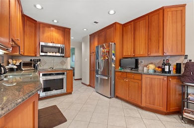 Kitchen featuring tasteful backsplash, brown cabinets, appliances with stainless steel finishes, dark stone countertops, and light tile patterned floors