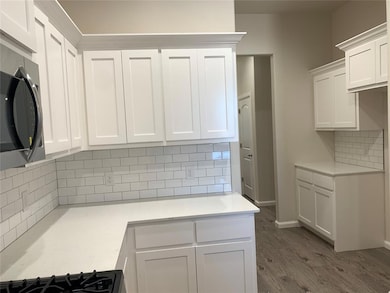 Kitchen featuring tasteful backsplash, white cabinetry, stainless steel microwave, wood finished floors, and light stone counters