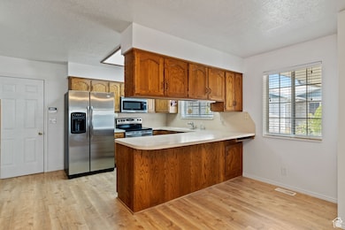 Kitchen featuring appliances with stainless steel finishes, brown cabinets, a peninsula, light wood finished floors, and light countertops